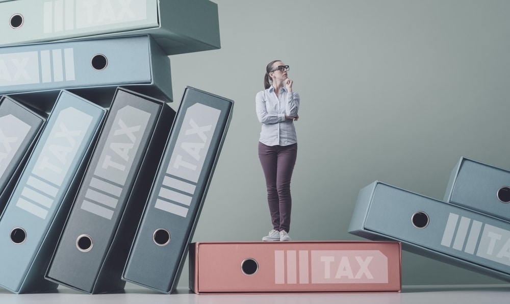 A woman wearing glasses with a questioning stance, standing on top of gigantic folders with tax labels